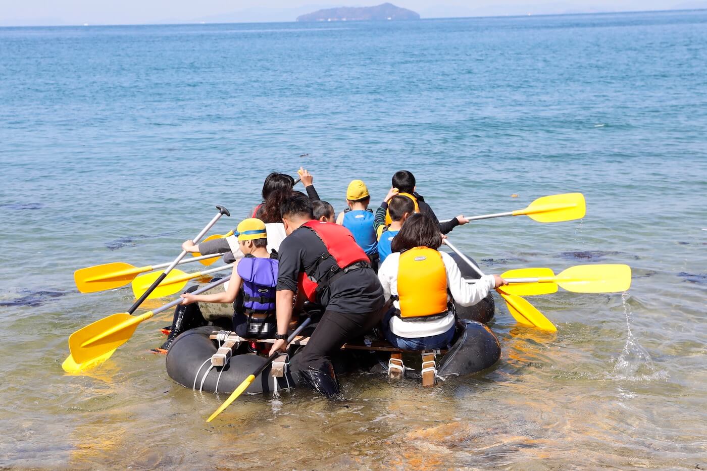 写真：一隻のイカダに乗って海へ漕ぎ出す複数の子どもと大人