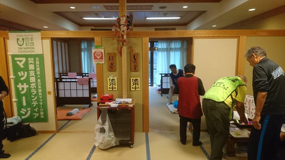 写真：温泉施設内にある畳の部屋の入り口付近で、マッサージの順番を待つ女性やスタッフの後ろ姿