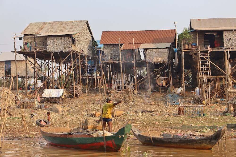 写真：川沿いに並ぶボロボロの家。川にはいまにも沈みそうな古い木の舟が浮かび、その上で仕事をする男性がいる