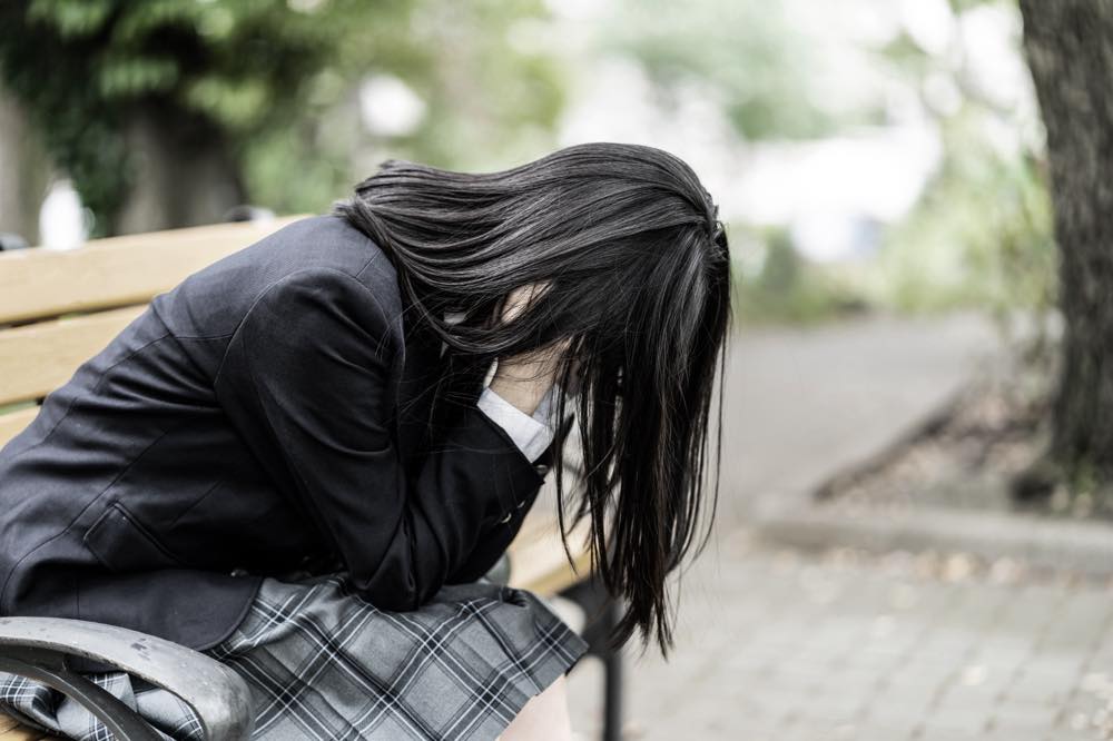 公園のベンチに座り、顔を手で覆ってうつむく女子学生。