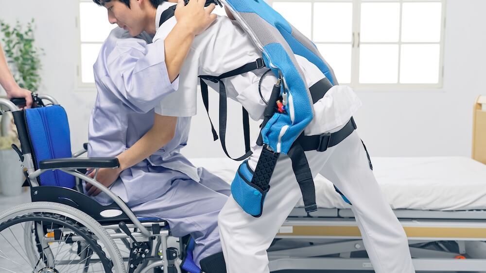 車椅子に乗った介護者が立つところをサポートする、パワーアシストを行う装着型の機器を着けた介助者