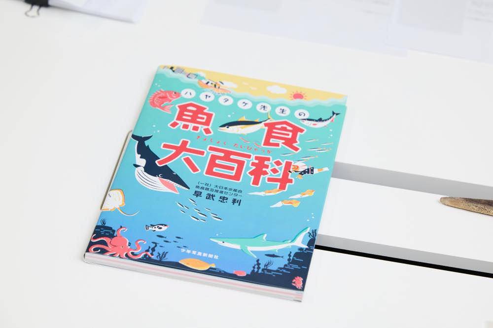 『ハヤタケ先生の魚食大百科』の表紙。中央に大きく『魚食大百科』、その上に『ハヤタケ先生の』と書かれている。表紙全体は海中と夕焼けの風景で構成され、さまざまな魚介類のイラストが描かれている。