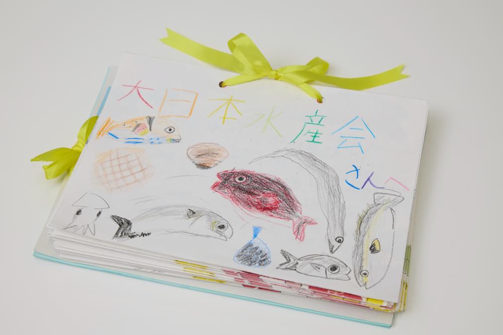 「『おさかな学習』に参加した子どもたちから届いた手紙の束。表紙には『大日本水産会さんへ』と書かれ、さまざまな魚のイラストが描かれている。表紙と手紙は黄色のリボンでまとめられている。」