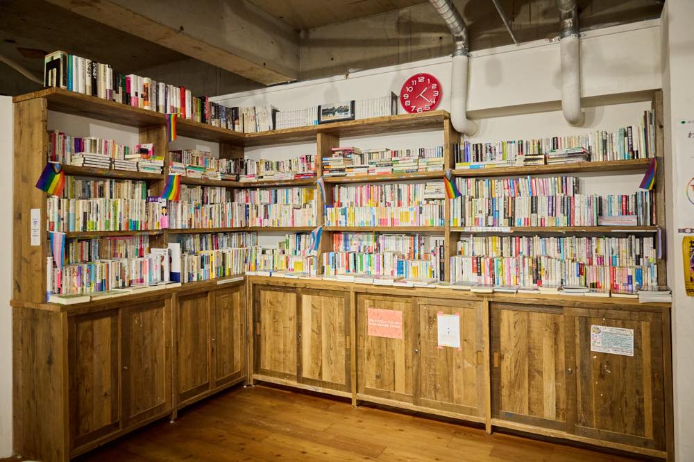 プライドハウス東京レガシー「LGBTQコミュニティ・アーカイブ＆ライブラリー1」の様子。
木製の本棚にLGBTQ+関連の本がぎっしりと並ぶ図書コーナー。棚にはレインボーフラッグが飾られ、温かみのある落ち着いた空間