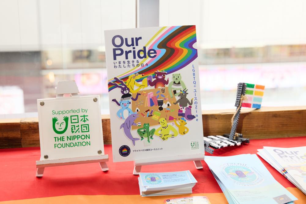 冊子『Our Pride』が棚の上に飾られている様子。

冊子の表紙には、プログレス・プライド・フラッグの11色（6色のレインボーカラーに加え、白・ピンク・水色のトランスジェンダーカラーと、茶色と黒の人種的マイノリティを表すカラー）が背景に描かれ、中央で動物たちがテーブルを囲んでいる。左上にはタイトル「Our Pride」とサブタイトル「いまを生きるわたしたちの日々」が記されている。
