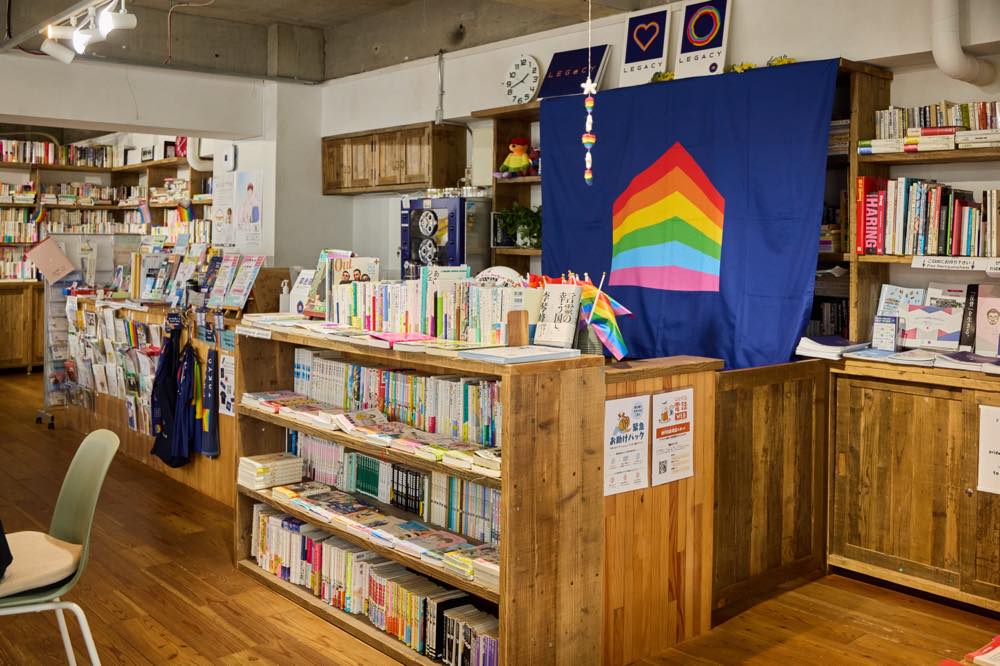 プライドハウス東京レガシーの「ミニキッチン」と「アーカイブ&ライブラリー2」の様子。「ミニキッチン」にはプライドハウス東京のロゴが印刷された大きな布が掲げられており、その周囲に「アーカイブ&ライブラリー2」が広がる。書籍や配布資料、プライドハウス東京のグッズが多数並んでいる。