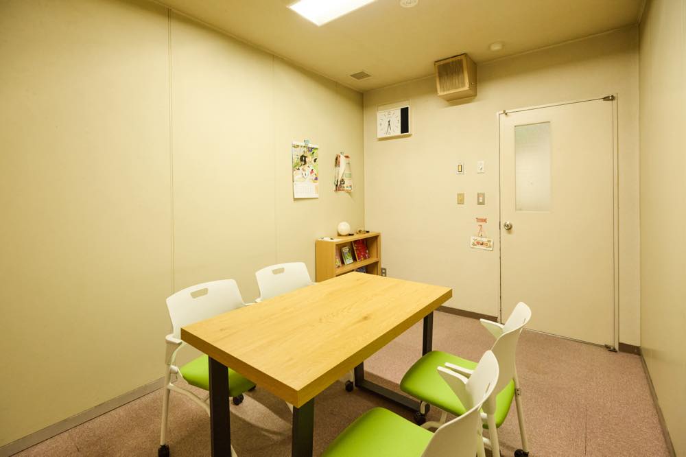 面談室の様子。中央に木製の机があり、向かい合う形で緑色のクッション付き椅子が2脚ずつ配置されている。ドア付近には本棚が置かれている。