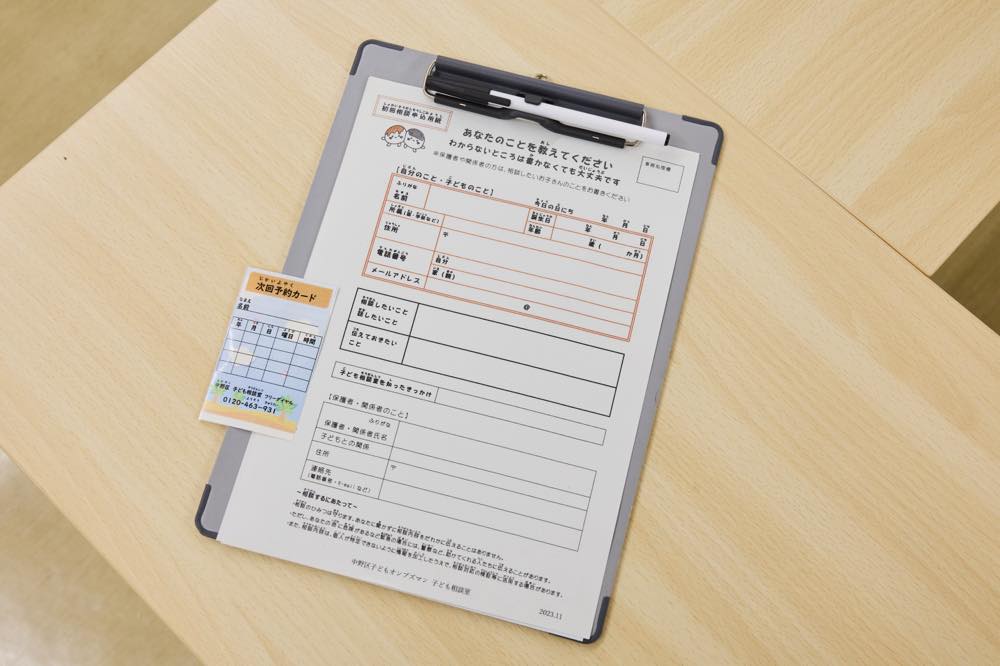 
記入用紙「あなたのことを教えてください（わからないところは書かなくても大丈夫です）」の写真。

用紙の上段は基本情報欄で、氏名・ふりがな、年齢、学校・学年、電話番号、メールアドレス、保護者（関係者）の氏名や続柄・連絡先などを書くマスが並ぶ。

その下に自由記述の枠があり、「相談したいこと」「話したいこと」「伝えておきたいこと」などを書く欄が続く。

さらに「子ども相談室を知ったきっかけ」を書く欄、保護者・関係者について（氏名・ふりがな、子どもとの関係、住所、連絡先 など）を書く欄が下部に配置されている。最下段には、記入上の注意や情報の取り扱いに関する説明文が小さく印字されている。

「次回予約カード」には、日付や時間を記入する表と、電話番号（0120-463-931）が印刷されている。