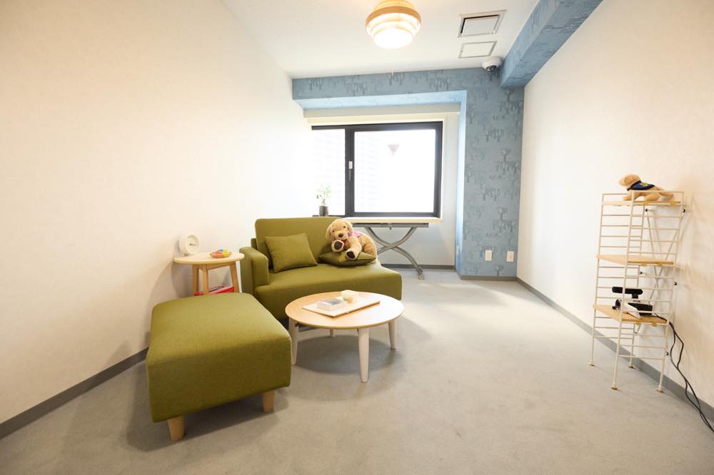 「つなっぐ」の司法面接室。窓から自然光が差し込み、温かな雰囲気が漂う部屋。緑色のソファが向かい合わせに二つ置かれ、その間には低い丸いテーブルがある。棚には、ソファとテーブルの方向を映すカメラが設置されている。