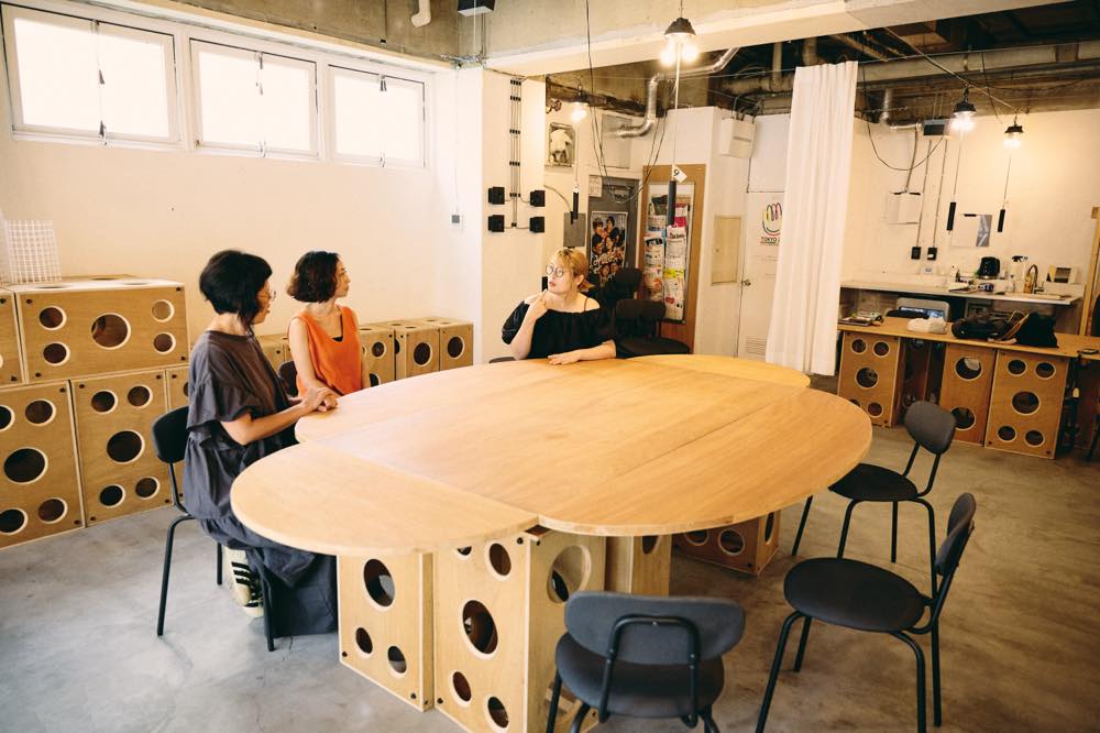 四角いテーブルの四辺に半円形の板を取り付けて作られた木製テーブルを囲み、3人が手話で会話している。