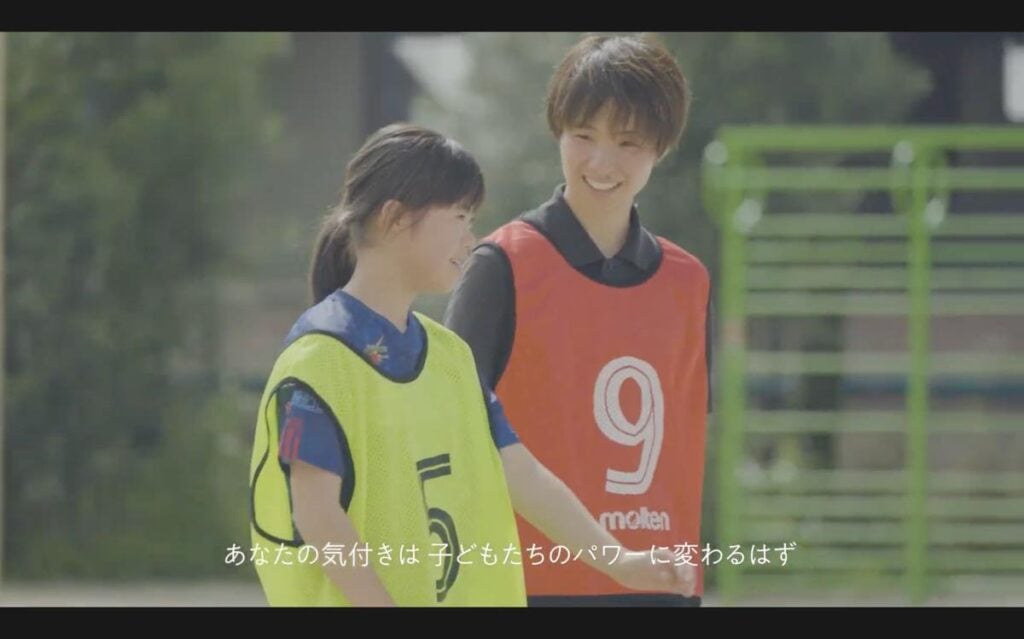 S.C.P.JapanのYouTubeで公開されている動画「スポーツにおけるセーフガーディング（コーチの声編）」のキャプチャー。井上さんと、黄色いゼッケンをつけた女の子が、並んで歩いている様子。画面下部には「あなたの気付きは 子どもたちのパワーに変わるはず」という白い字幕が表示されている。