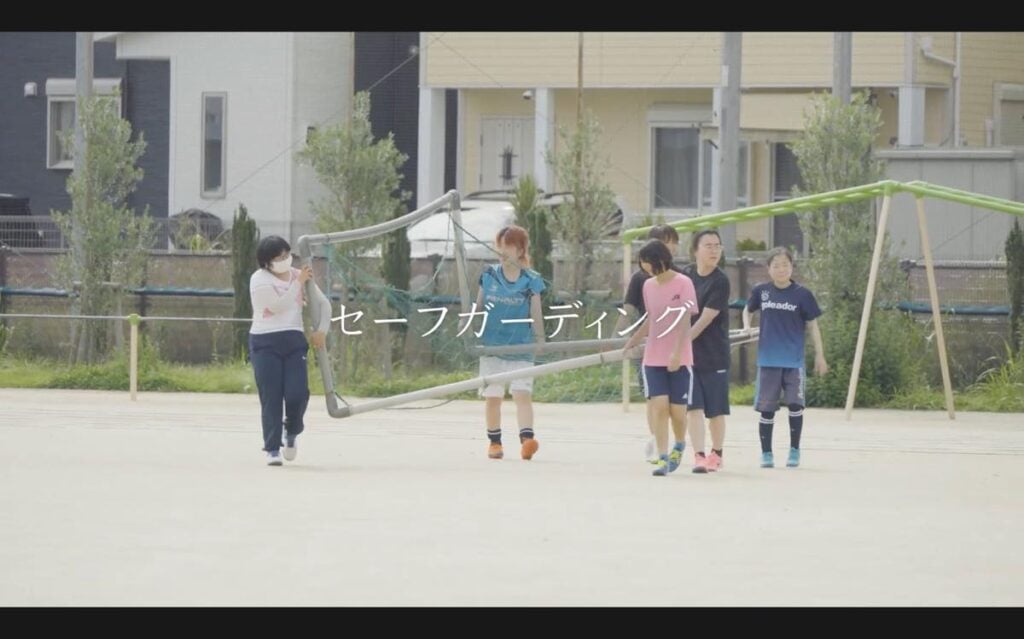 S.C.P.JapanのYouTubeで公開されている動画「スポーツにおけるセーフガーディング（コーチの声編）」のキャプチャー。屋外のグラウンドで、子どもとスタッフがサッカーゴールのフレームを運んでいる様子。画面中央には大きく「セーフガーディング」という白い文字が書かれている。