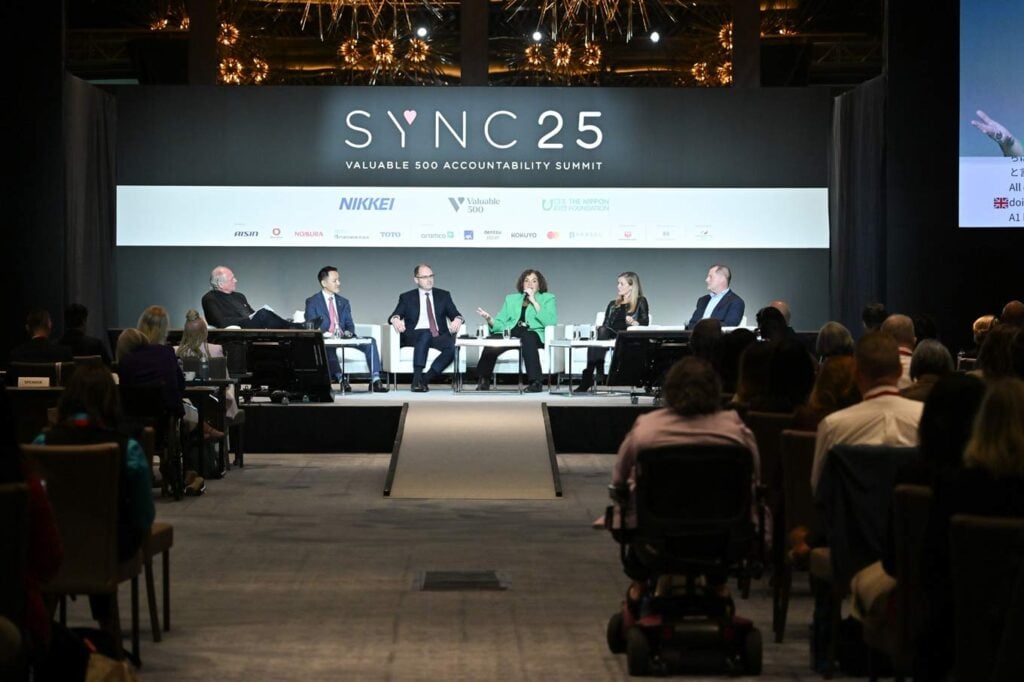 写真：SYNC25アカウンタビリティ・サミットで、世界的大企業の経営幹部複数名がステージ上で障害者インクルージョンについてパネルディスカッションを行っている様子。客席には車椅子利用者を含む参加者が座っている。）