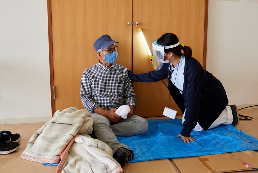 避難所のような室内で、青いシートと段ボールの上に座る高齢男性と、その肩に手を添えて話しかける女性スタッフの写